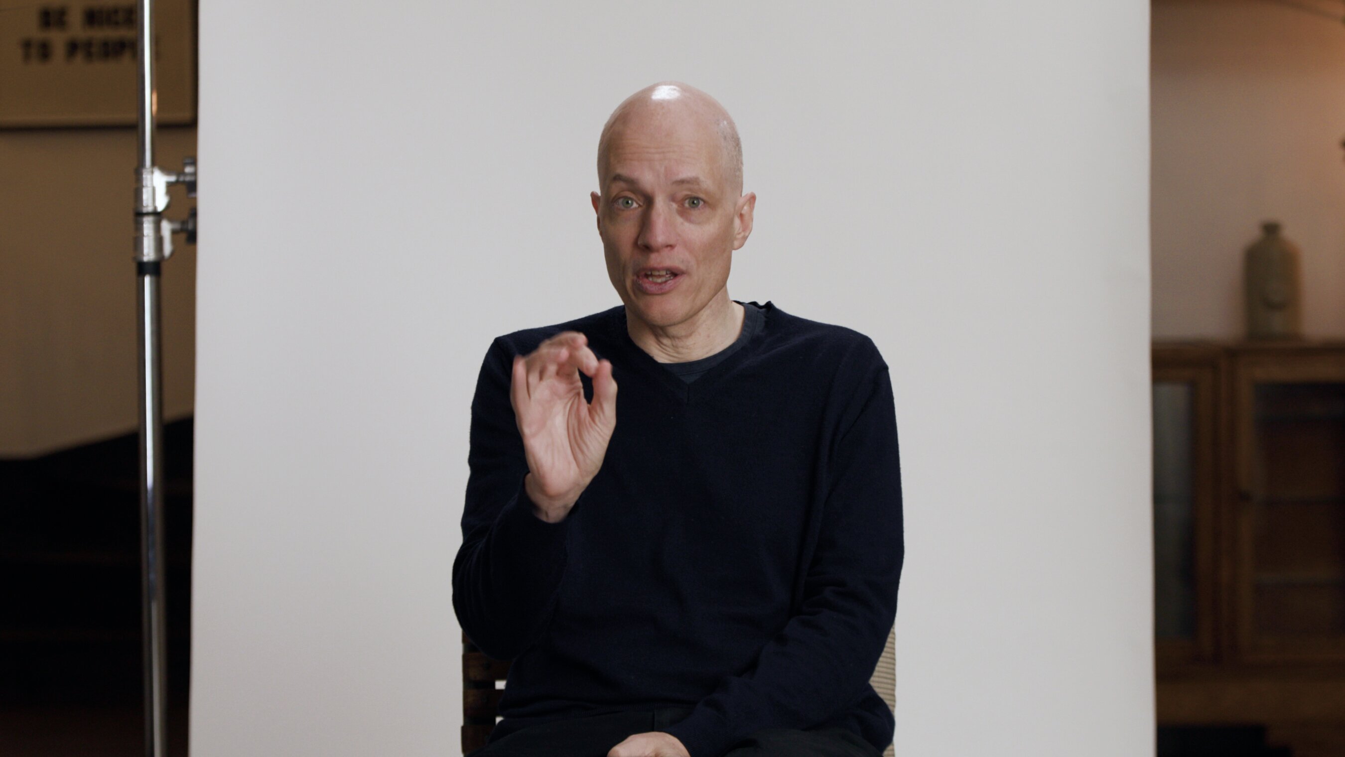 A bald man wearing a black sweater sits against a plain light background, gesturing with his right hand while looking at the camera.