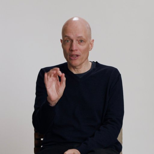 A bald man wearing a black sweater sits against a plain light background, gesturing with his right hand while looking at the camera.