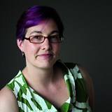 A person with short purple hair, glasses, and a patterned green and white sleeveless top poses against a dark background.
