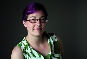 A person with short purple hair, glasses, and a patterned green and white sleeveless top poses against a dark background.
