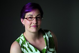 A person with short purple hair, glasses, and a patterned green and white sleeveless top poses against a dark background.