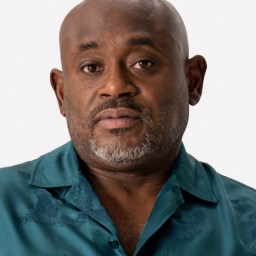 A middle-aged man with a bald head, trimmed gray beard, and teal button-up shirt looks directly at the camera against a plain background.