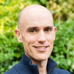 A man with a shaved head, light skin, and a dark zip-up sweater smiles at the camera with greenery in the background.