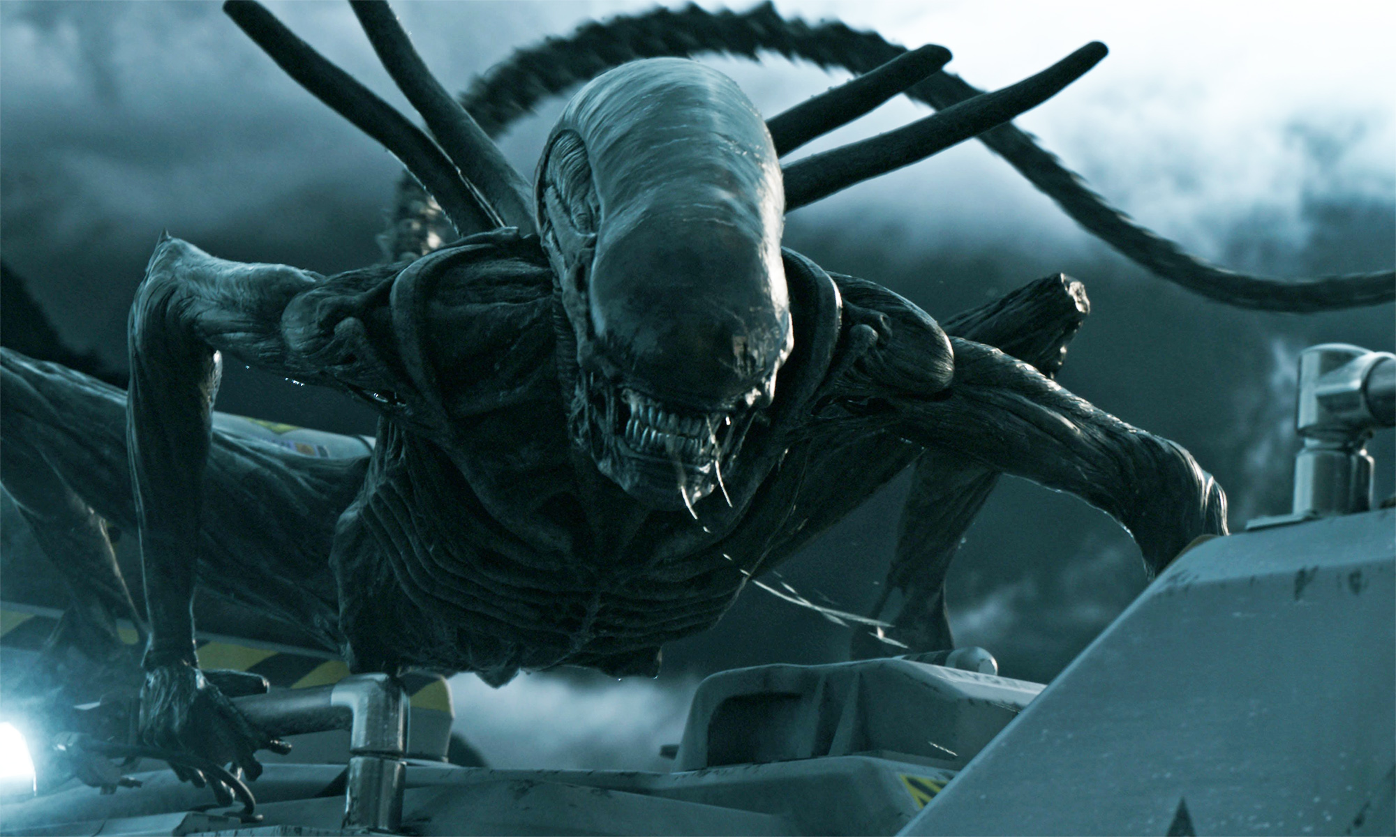 A large, dark, menacing alien creature with an elongated head and sharp teeth crouches on a metallic surface under a cloudy sky, embodying the fearsome presence often associated with aliens.