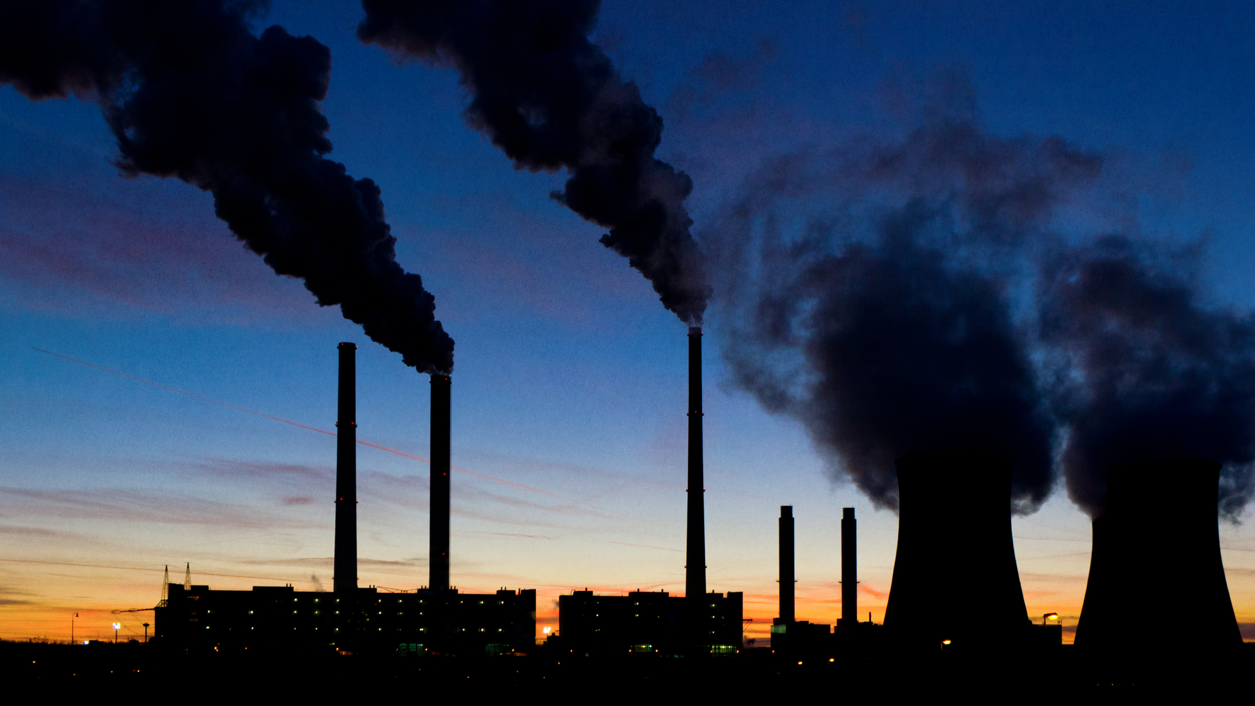 Industrial facility with multiple smokestacks and cooling towers emitting smoke or steam against a dusk sky.