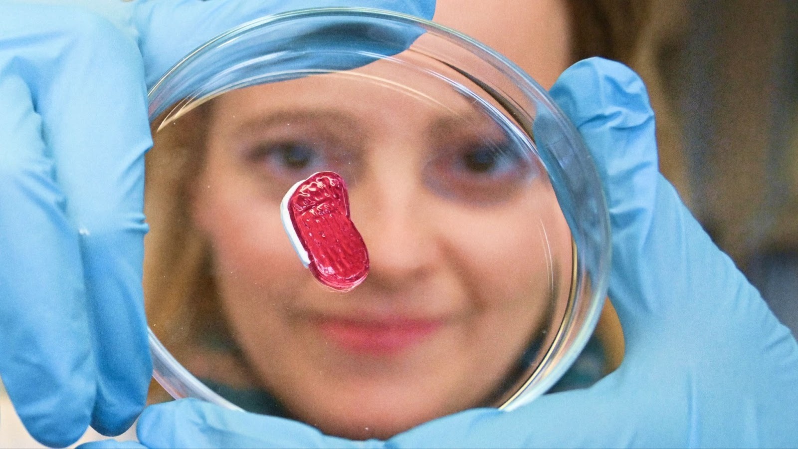 A person wearing blue gloves holds a petri dish containing a small pink object shaped like a human foot.