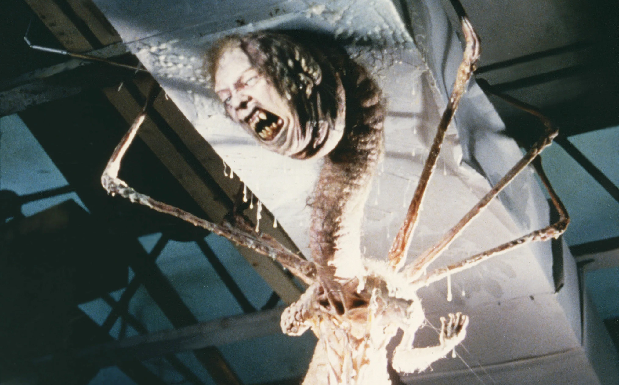 A creature with a distorted human face and long, spindly limbs&mdash;resembling twisted aliens&mdash;emerges from a ceiling covered in slime and organic material.