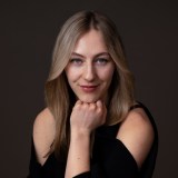 Woman with long blonde hair and blue eyes poses with her chin resting on her fist, wearing a black top against a dark background.