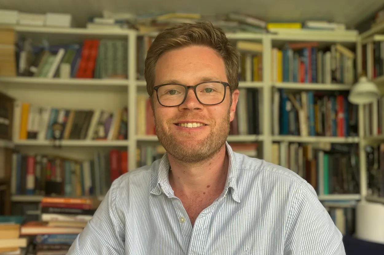 A man with glasses and a light blue striped shirt is smiling in front of a bookshelf filled with books.