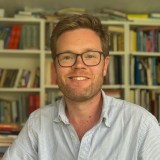 A man with glasses and a light blue striped shirt is smiling in front of a bookshelf filled with books.