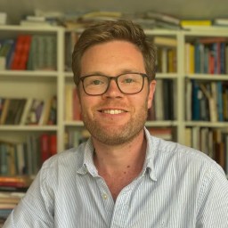 A man with glasses and a light blue striped shirt is smiling in front of a bookshelf filled with books.