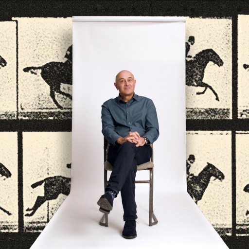 A man sits on a chair against a white backdrop, with a background featuring repeated vintage images of a person riding a horse.