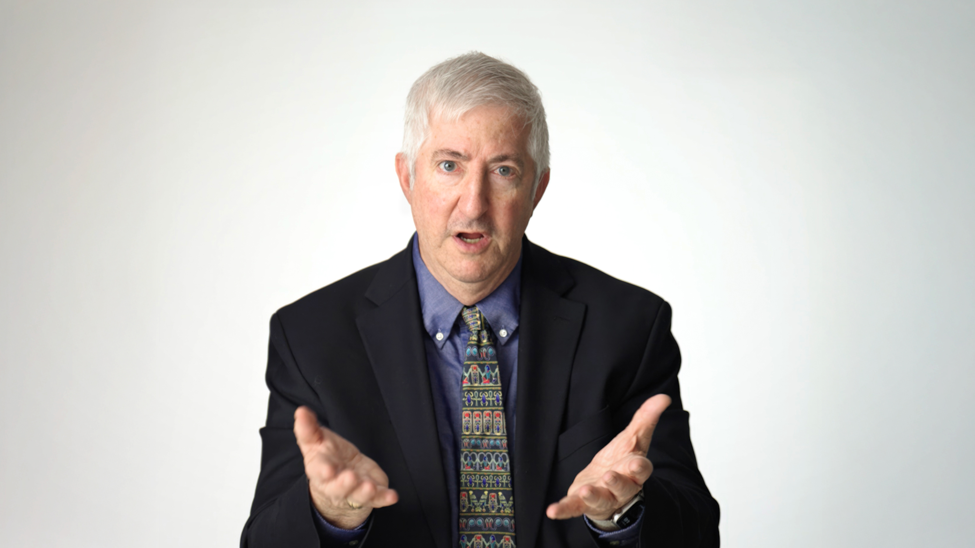 Older man with gray hair wearing a dark suit, patterned tie, and blue shirt, gesturing with both hands, seated against a plain white background.