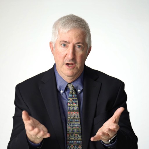 Older man with gray hair wearing a dark suit, patterned tie, and blue shirt, gesturing with both hands, seated against a plain white background.