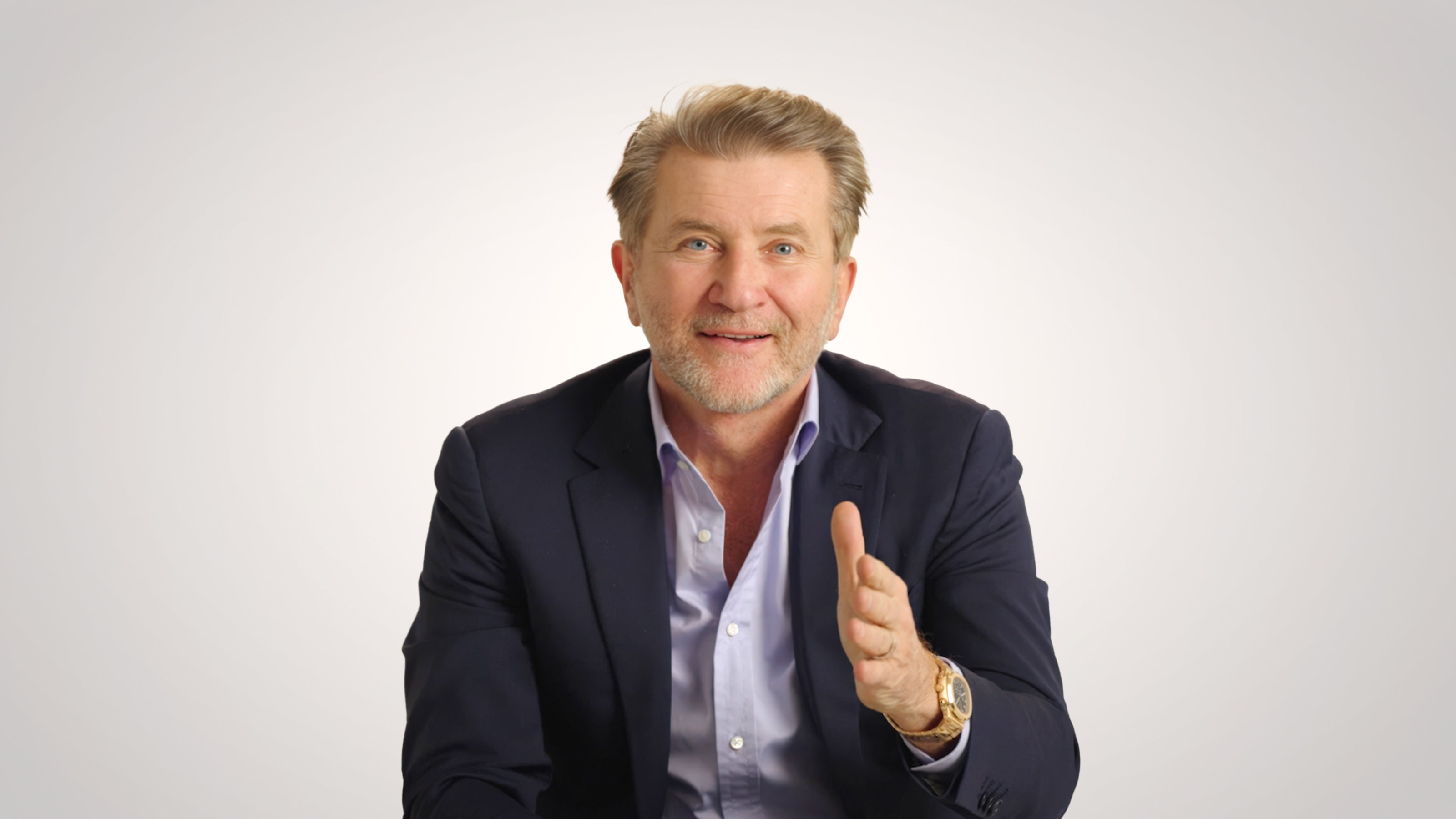 A middle-aged man in a navy suit and light blue shirt gestures with his right hand while sitting against a plain light background.