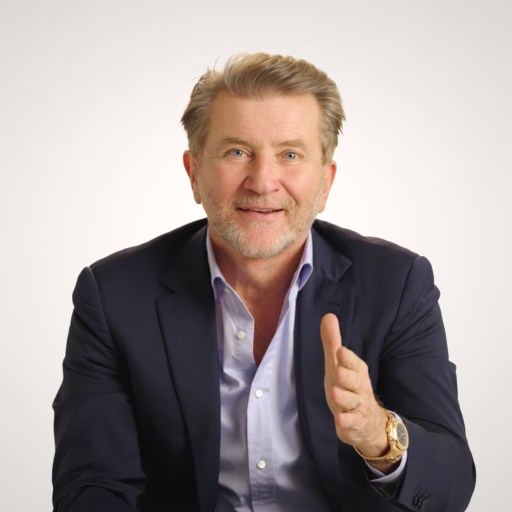 A middle-aged man in a navy suit and light blue shirt gestures with his right hand while sitting against a plain light background.