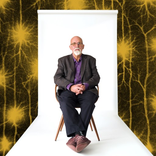 An older man with a beard sits on a chair in front of a white backdrop, with yellow neuron-like patterns on a black background surrounding the scene.