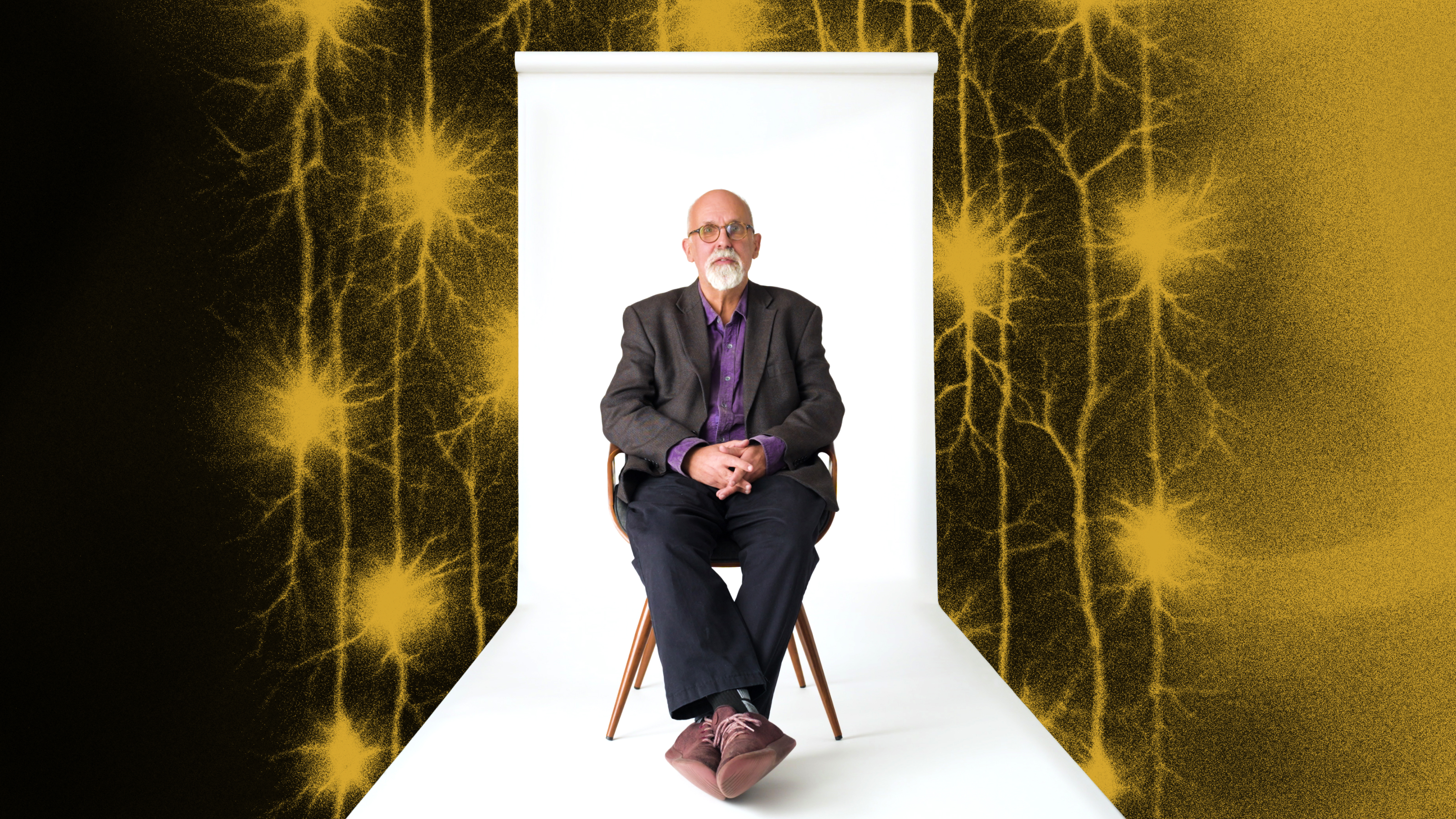 An older man with a beard sits on a chair in front of a white backdrop, with yellow neuron-like patterns on a black background surrounding the scene.