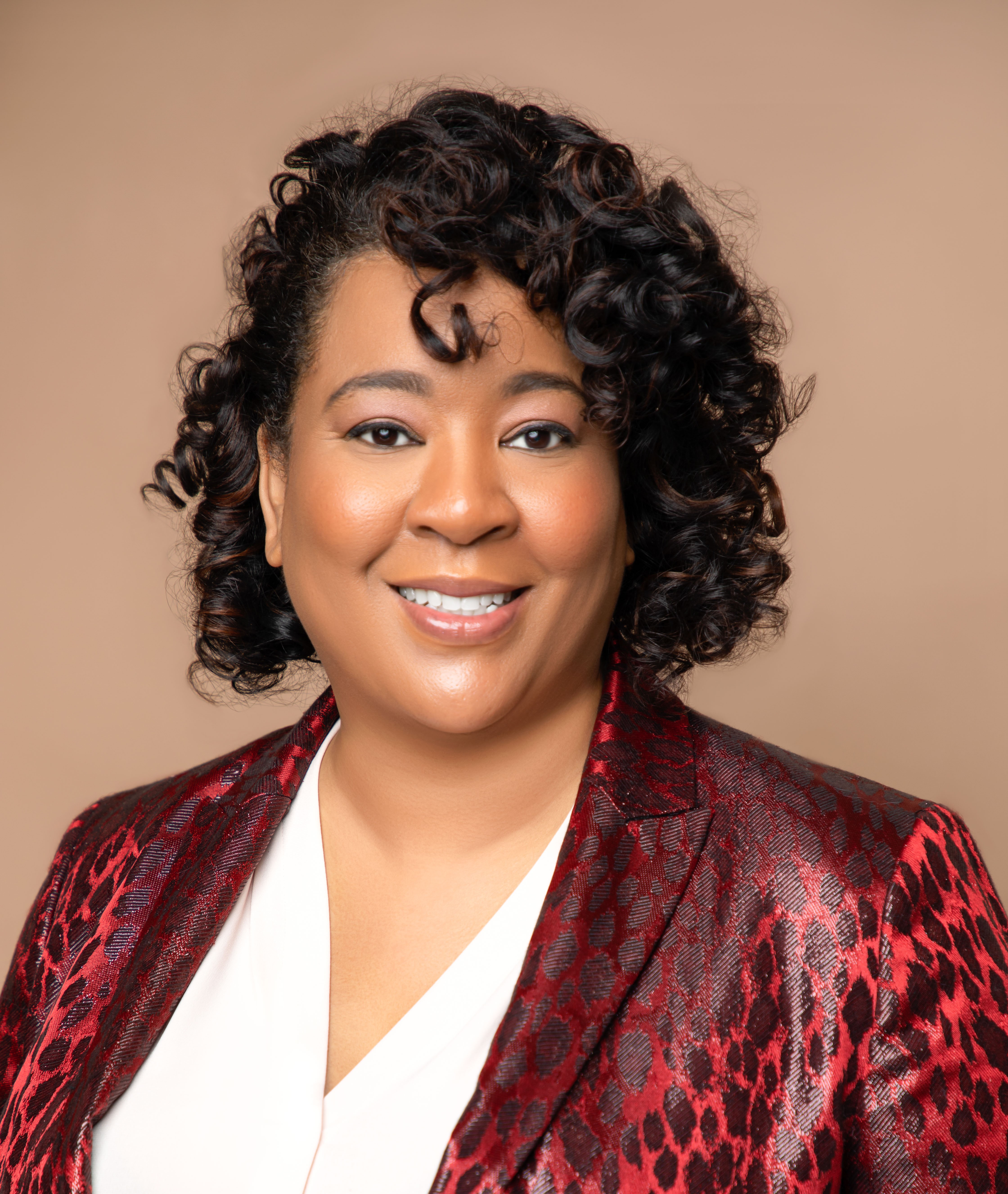 A woman with curly hair wearing a red patterned blazer and a white blouse, smiling against a plain beige background.