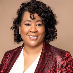 A woman with curly hair wearing a red patterned blazer and a white blouse, smiling against a plain beige background.