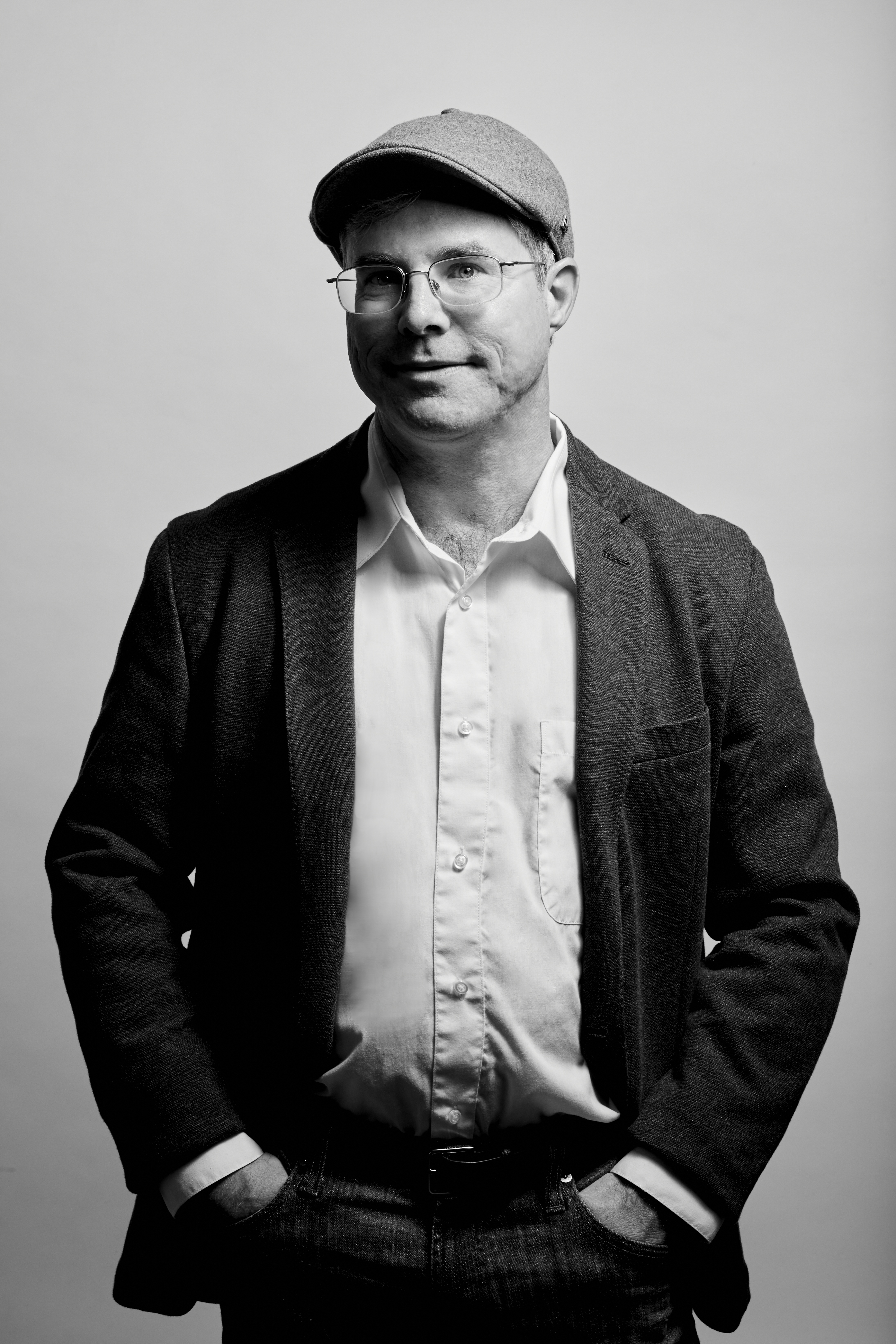A man wearing a flat cap, glasses, blazer, and button-up shirt poses with hands in pockets against a plain background in a black and white photo.