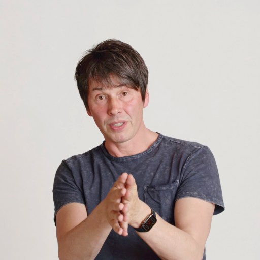 A man with short dark hair, wearing a dark t-shirt and smartwatch, gestures with his hands while standing in front of a plain white background.