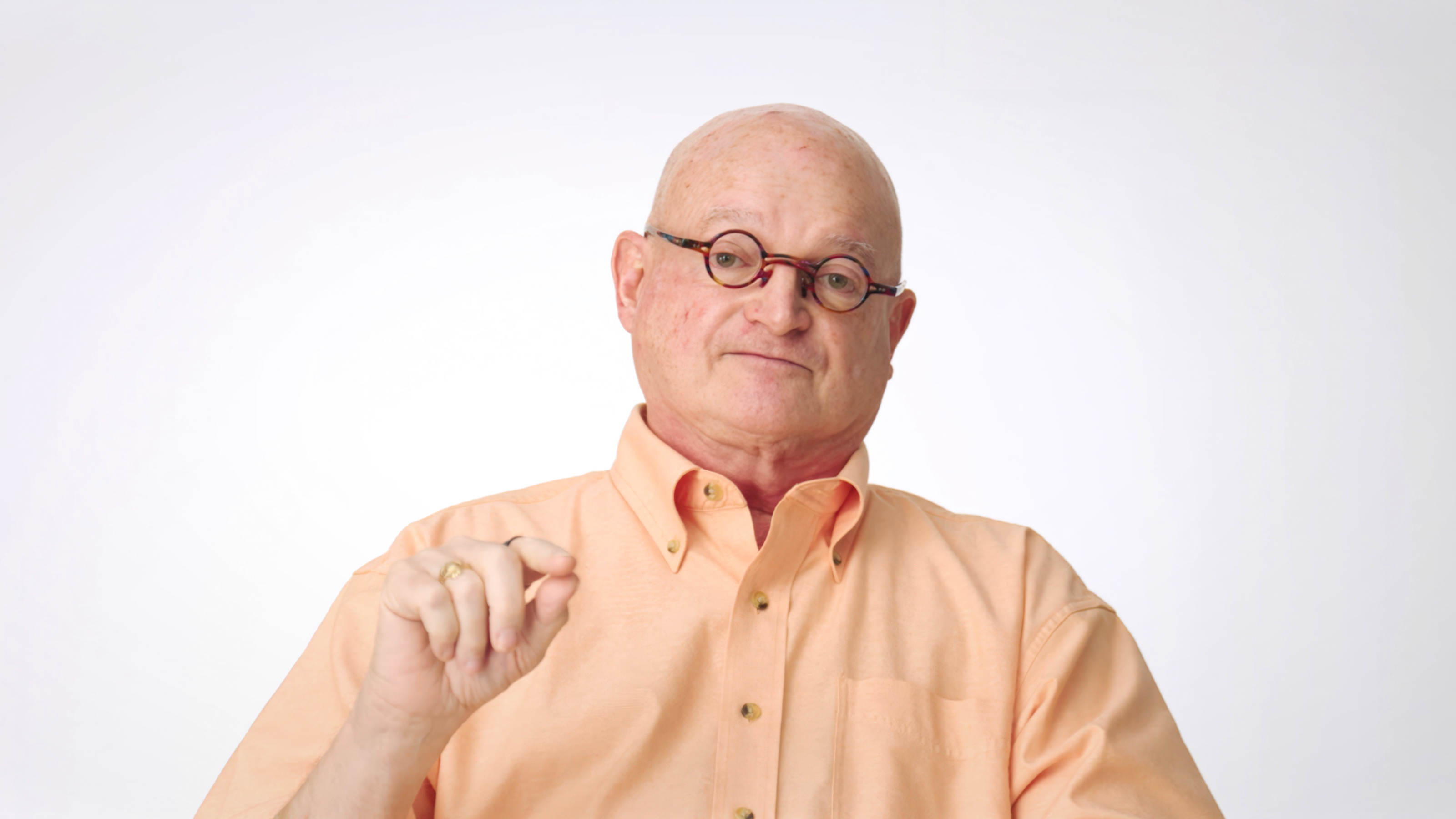 A bald man wearing glasses and a peach-colored button-up shirt sits facing the camera against a plain white background, holding his right hand slightly raised.