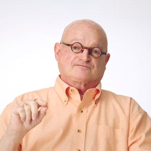 A bald man wearing glasses and a peach-colored button-up shirt sits facing the camera against a plain white background, holding his right hand slightly raised.