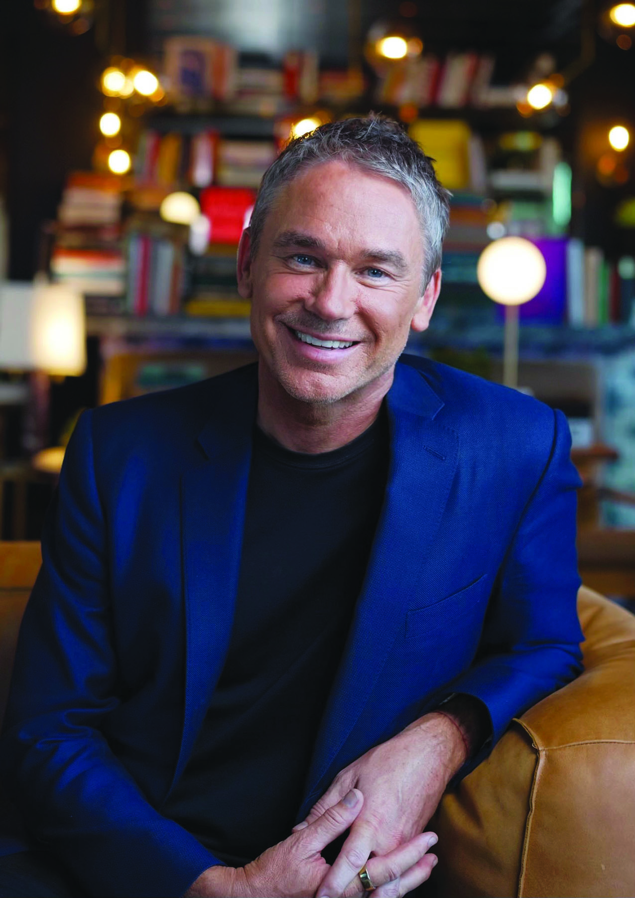 A man with short gray hair wearing a blue blazer and black shirt sits on a brown couch, smiling, with bookshelves and warm lighting in the background.