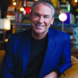 A man with short gray hair wearing a blue blazer and black shirt sits on a brown couch, smiling, with bookshelves and warm lighting in the background.