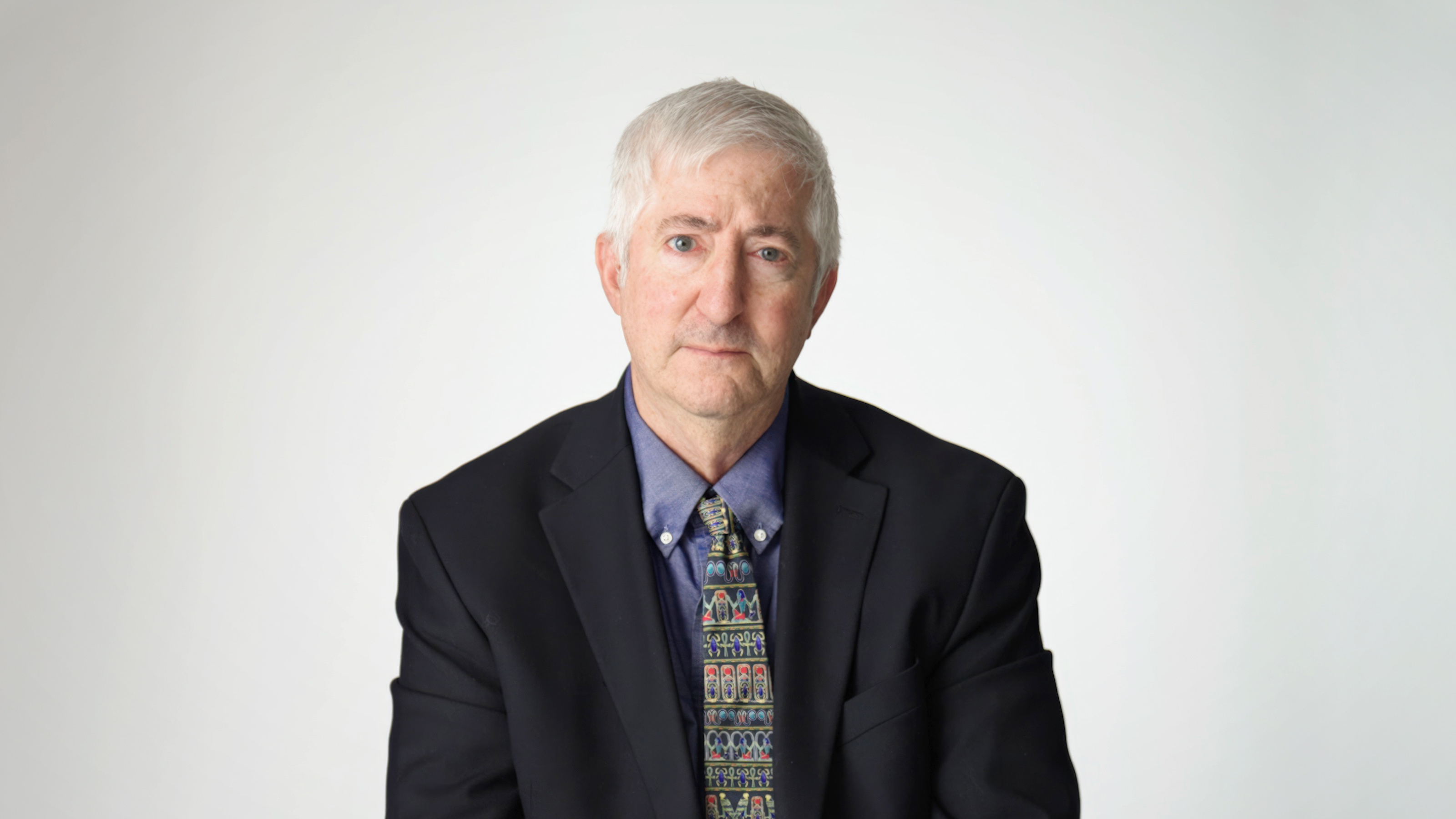 An older man with gray hair wearing a dark suit, blue shirt, and patterned tie, sitting against a plain light background.