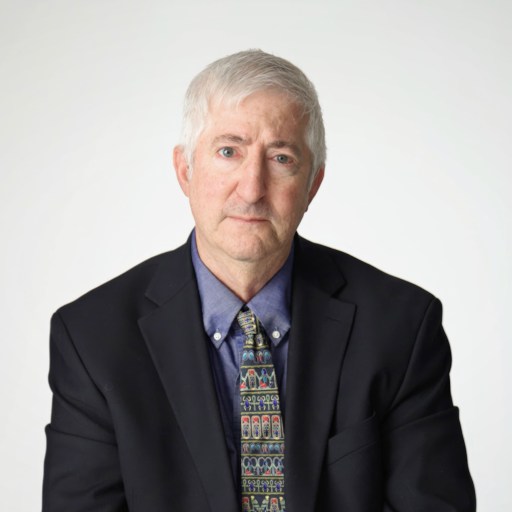 An older man with gray hair wearing a dark suit, blue shirt, and patterned tie, sitting against a plain light background.