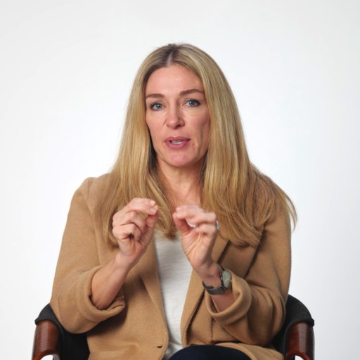A woman with long blonde hair sits on a chair against a plain white background, wearing a tan jacket and gesturing with both hands while speaking.