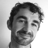 A man with curly hair and facial stubble smiles slightly, facing the camera against a plain light background.