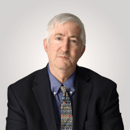 An older man with short gray hair, wearing a dark suit jacket, blue shirt, and patterned tie, sits against a plain light background.