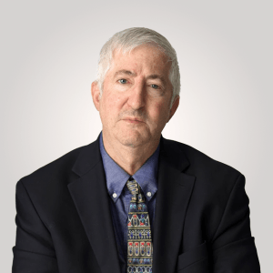 An older man with short gray hair, wearing a dark suit jacket, blue shirt, and patterned tie, sits against a plain light background.
