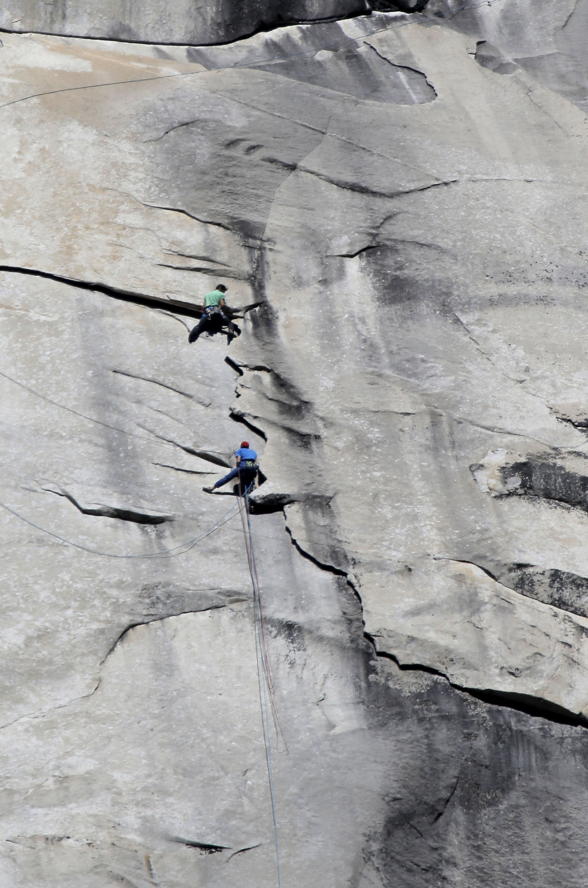 Two rock climbers, inspired by legends like Tommy Caldwell, ascend a steep, gray granite wall, attached to ropes and wearing helmets and harnesses.