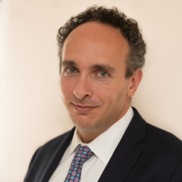 A man with short curly hair wearing a dark suit, white shirt, and patterned tie, standing against a plain light-colored background.