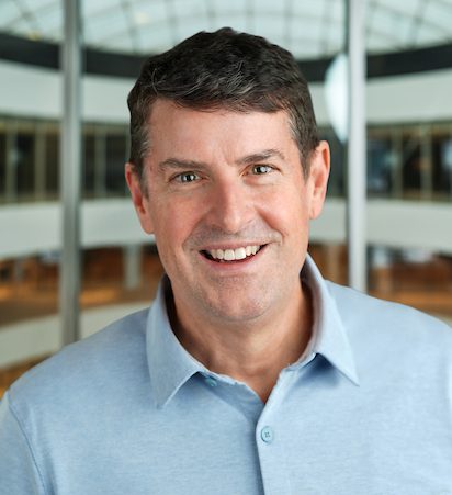 A man with short dark hair, wearing a light blue collared shirt, smiles in an indoor setting with a curved glass ceiling and blurred background.