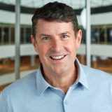 A man with short dark hair, wearing a light blue collared shirt, smiles in an indoor setting with a curved glass ceiling and blurred background.