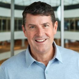 A man with short dark hair, wearing a light blue collared shirt, smiles in an indoor setting with a curved glass ceiling and blurred background.