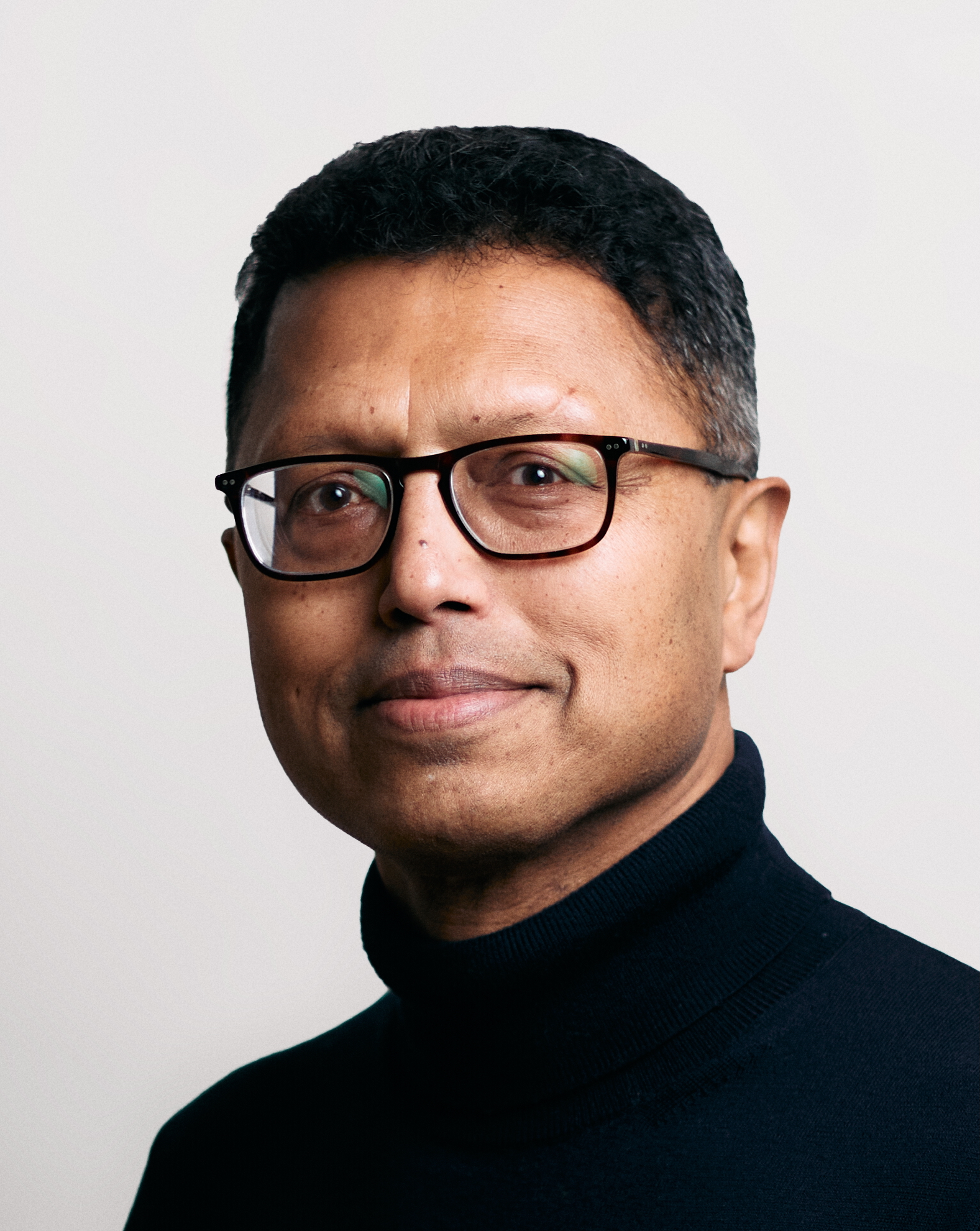 A man wearing glasses and a black turtleneck sweater poses for a portrait against a plain light background.
