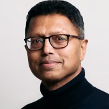 A man wearing glasses and a black turtleneck sweater poses for a portrait against a plain light background.