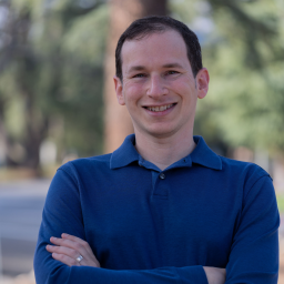 A man wearing a blue collared shirt stands outdoors with his arms crossed, smiling at the camera. Trees and a road are visible in the blurred background.