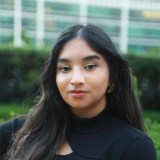 A woman with long dark hair wearing a black top poses outdoors in front of greenery and a blurred glass building.