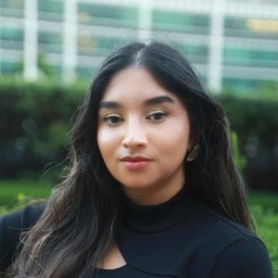 A woman with long dark hair wearing a black top poses outdoors in front of greenery and a blurred glass building.