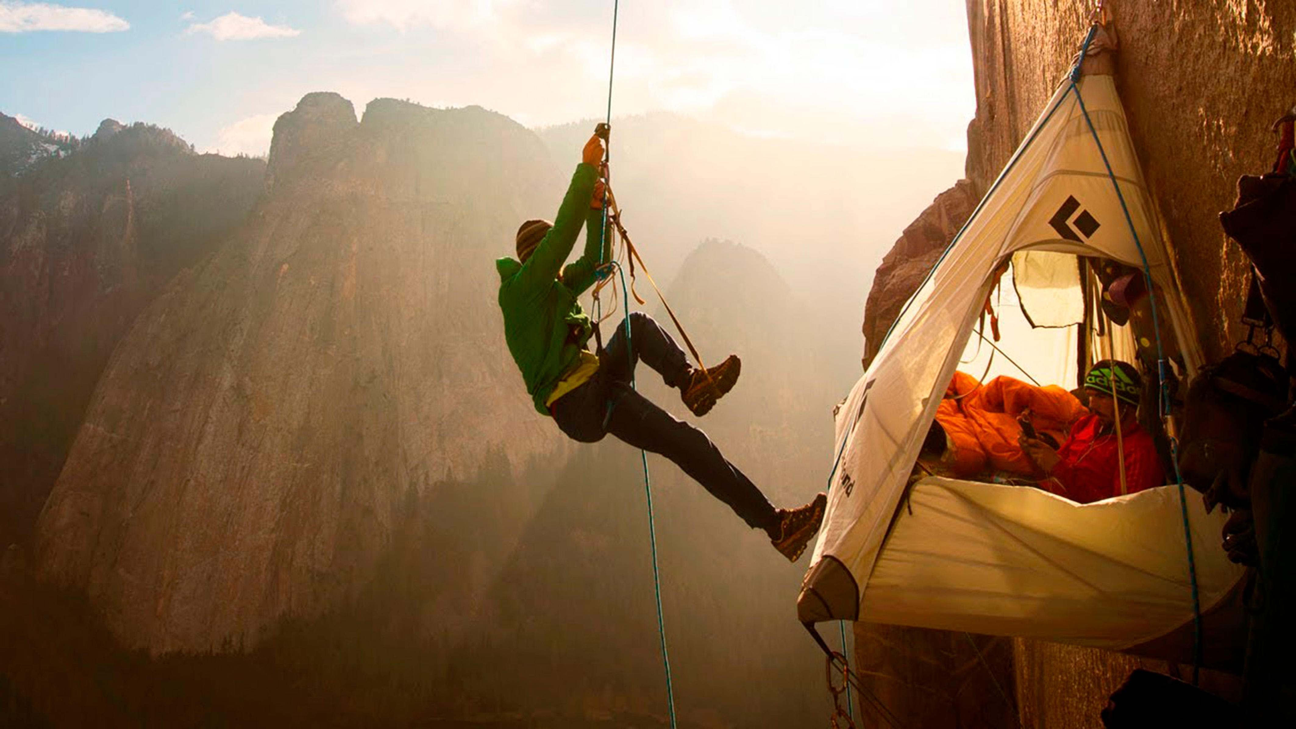 Climber Tommy Caldwell in a green jacket rappels beside a hanging tent attached to a cliff face, with another person inside and mountains in the background.