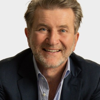 A middle-aged man with light brown hair and a trimmed beard smiles at the camera, wearing a dark blazer over a light blue shirt against a plain background.