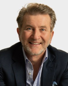 A middle-aged man with light brown hair and a trimmed beard smiles at the camera, wearing a dark blazer over a light blue shirt against a plain background.