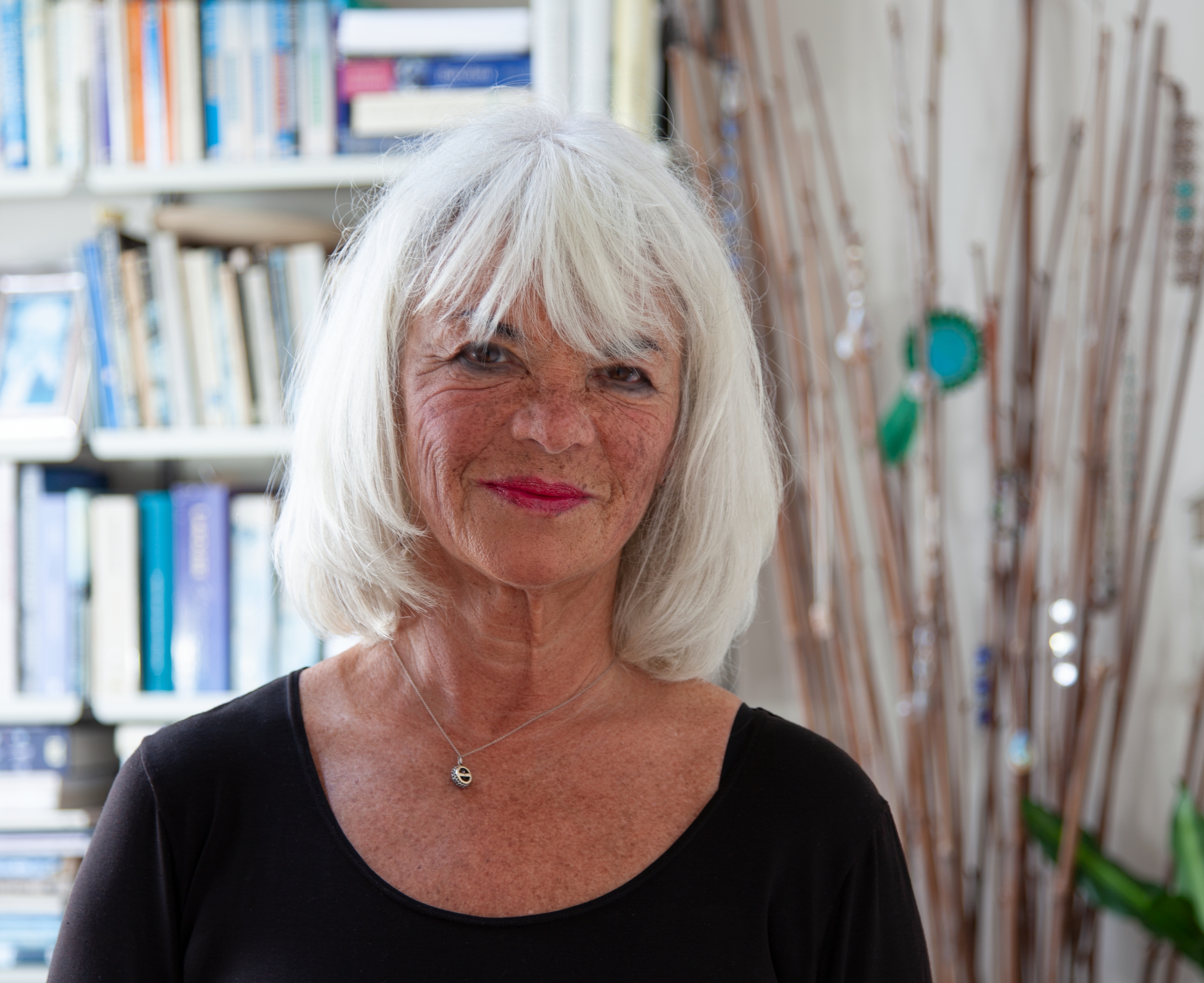 An older woman with white hair and a black top stands indoors in front of a bookshelf and decorative branches, smiling at the camera.